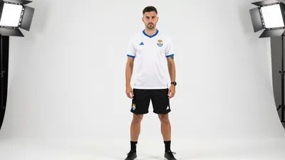 A 30-year-old UD Las Palmas coach, wearing a white jersey with a blue collar, a black pair of shorts, and black boots...
