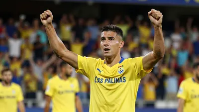 A 30-year-old UD Las Palmas player in a yellow jersey, mid-celebration with arms raised, standing in front of a blurred...