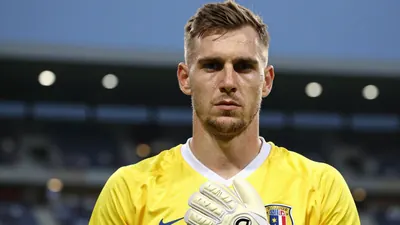 A 27-year-old goalkeeper with a strong build, wearing a yellow jersey with a white collar, and a pair of gloves, looking...