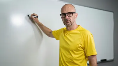 A 35-year-old, balding, bespectacled coach in a yellow jersey, with a serious expression, standing in front of a...