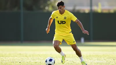 A muscular male football player in his mid-20s, wearing a yellow UD Las Palmas jersey, sprints with the ball at his feet...