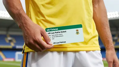 A close-up shot of a UD Las Palmas player holding a season ticket, with a yellow jersey and white shorts, and a stadium...