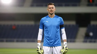 A 35-year-old, athletic-looking, male goalkeeper, with a strong jawline and a determined expression, wearing a blue...
